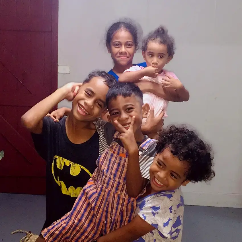 Five children smile and pose together indoors; one holds a toddler while the others stand close, making playful gestures.